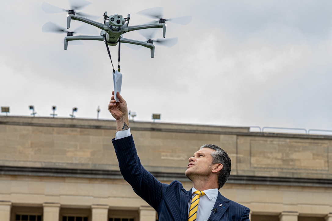 A man in a suit pulls a piece of paper attached to a drone hovering in the air during a cloudy day. A building with columns is in the background.