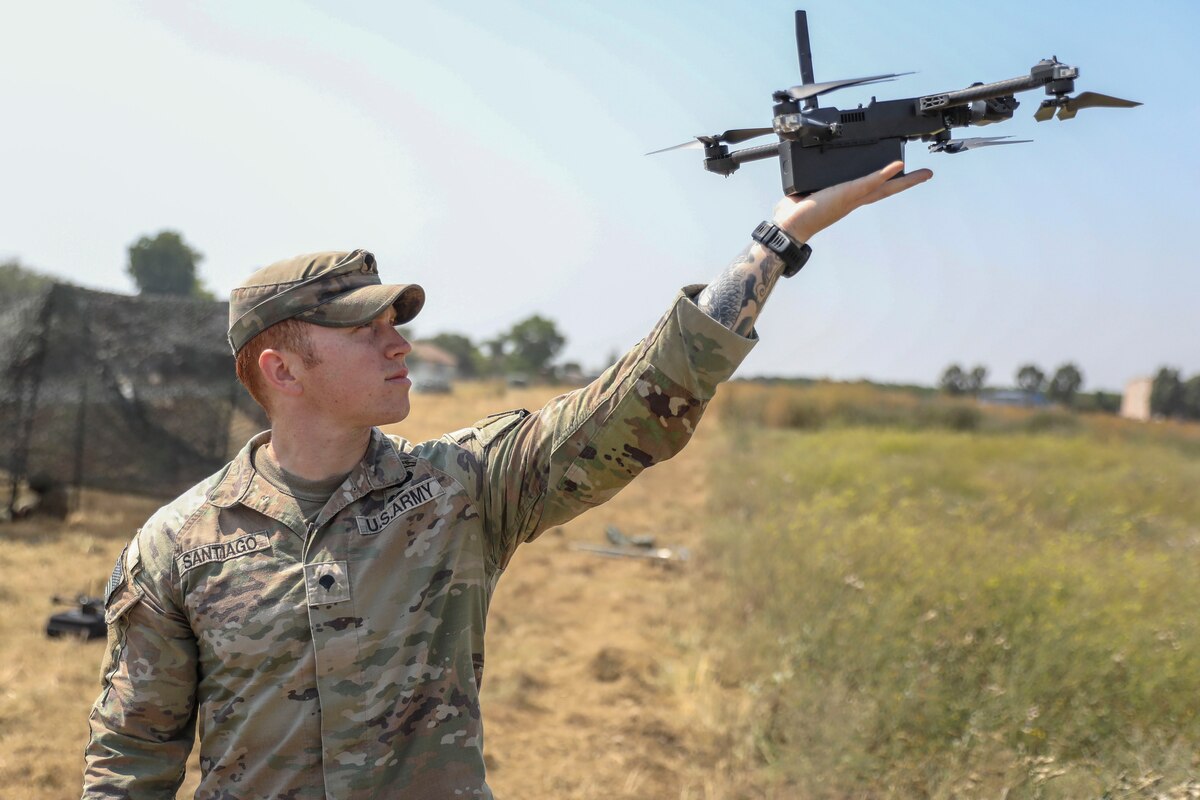 Outdoors in a field, a soldier in a camouflage uniform holds a small drone aircraft in his outstretched left arm.