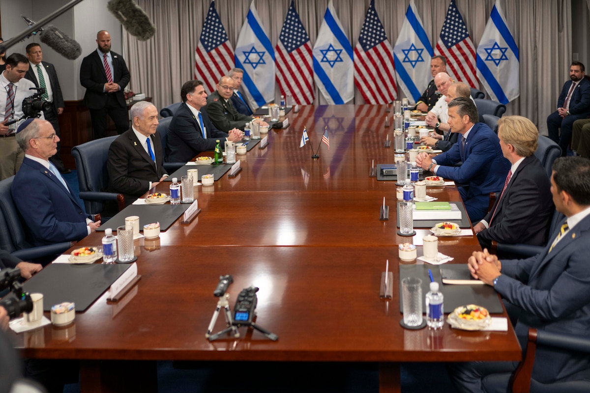 Nearly a dozen individuals in business attire sit around a large wooden table. In the rear are both U.S. and Israeli flags against the wall. Above their heads are boom microphones held by journalists.