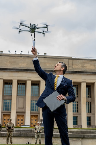 A man in a business suit extends his right arm into the air to retrieve a folded piece of paper hanging beneath a small, hovering drone. In the rear is a large building and two service members in camouflage uniforms holding remote control devices.