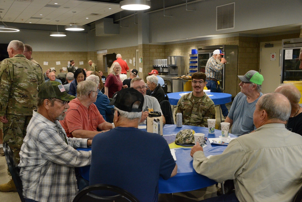 Past and present members of the 185th Air Refueling Wing sit and stand in a dining facility, speaking, eating and drinking coffee