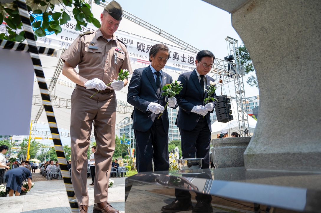 Three people wearing white gloves and holding flowers bow their heads toward an outdoor monument as others sit or stand in the background.