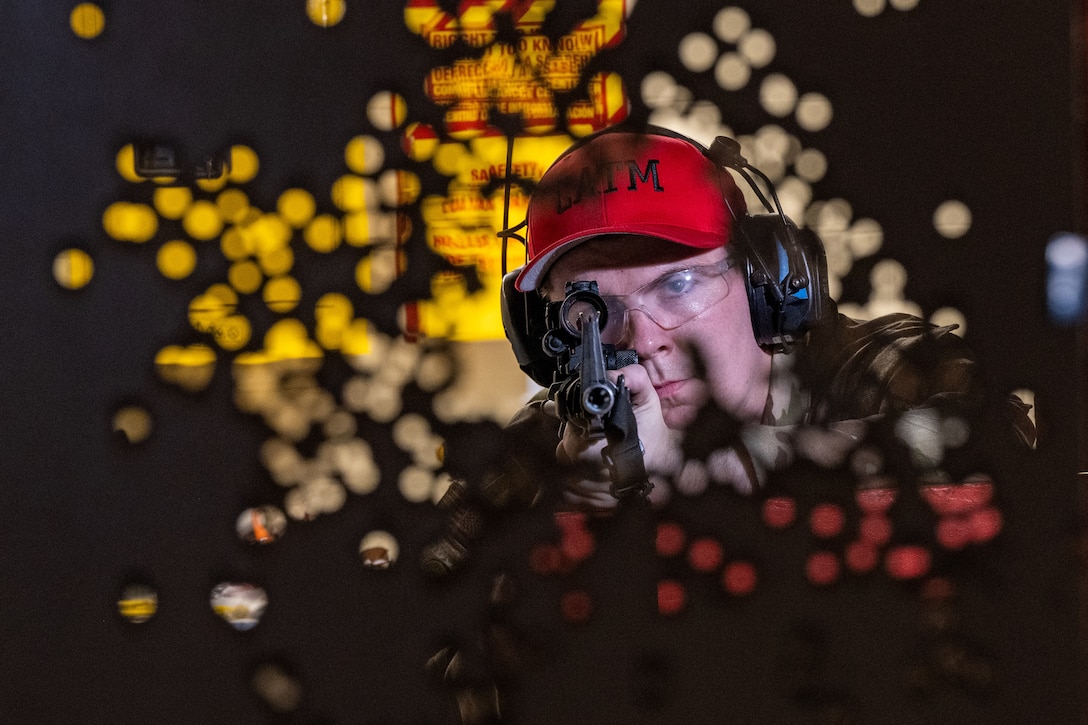 An airman is seen through the back side of a shooting target, wearing a red cap, ear protection and looking through the scope of a rifle.