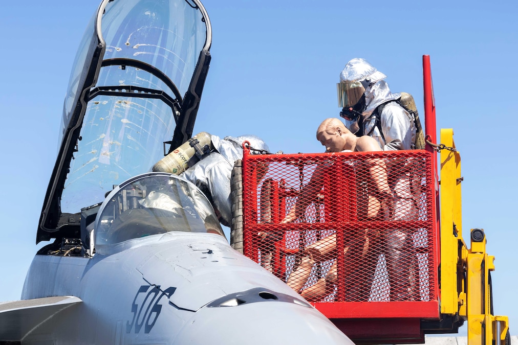 Two sailors in firefighting gear, one standing in the cockpit of a jet and one on an elevated platform next to the jet, pull a mannequin away from the aircraft.