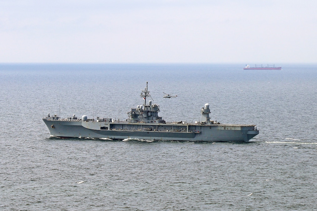 A ship cruises the sea with a helicopter flying nearby and another ship in the distance.