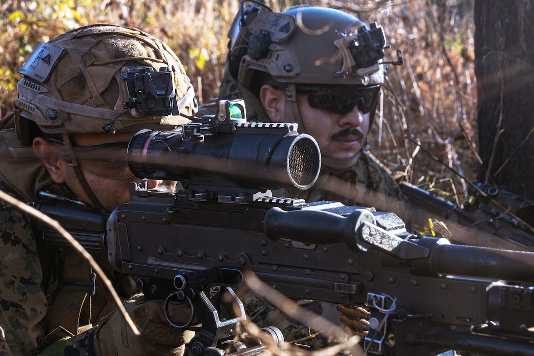 Two Marines in combat gear, one peering through the scope of a large machine gun, lie in a wooded area.