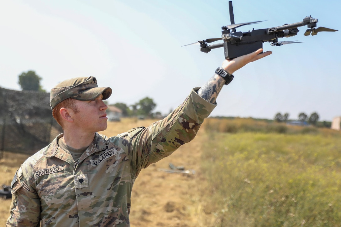 A soldier standing in a grassy field holds a drone aloft in one hand with a blue sky in the background.