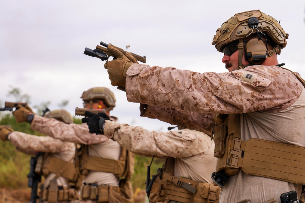A Marine wearing a helmet, sunglasses and gloves fires a pistol along with others lined in a row outdoors.
