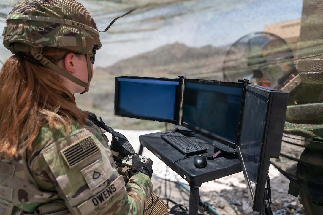A soldier sitting under netting outdoors holds a device to operate a surveillance system with three screens and a keyboard on a table in front of the soldier.