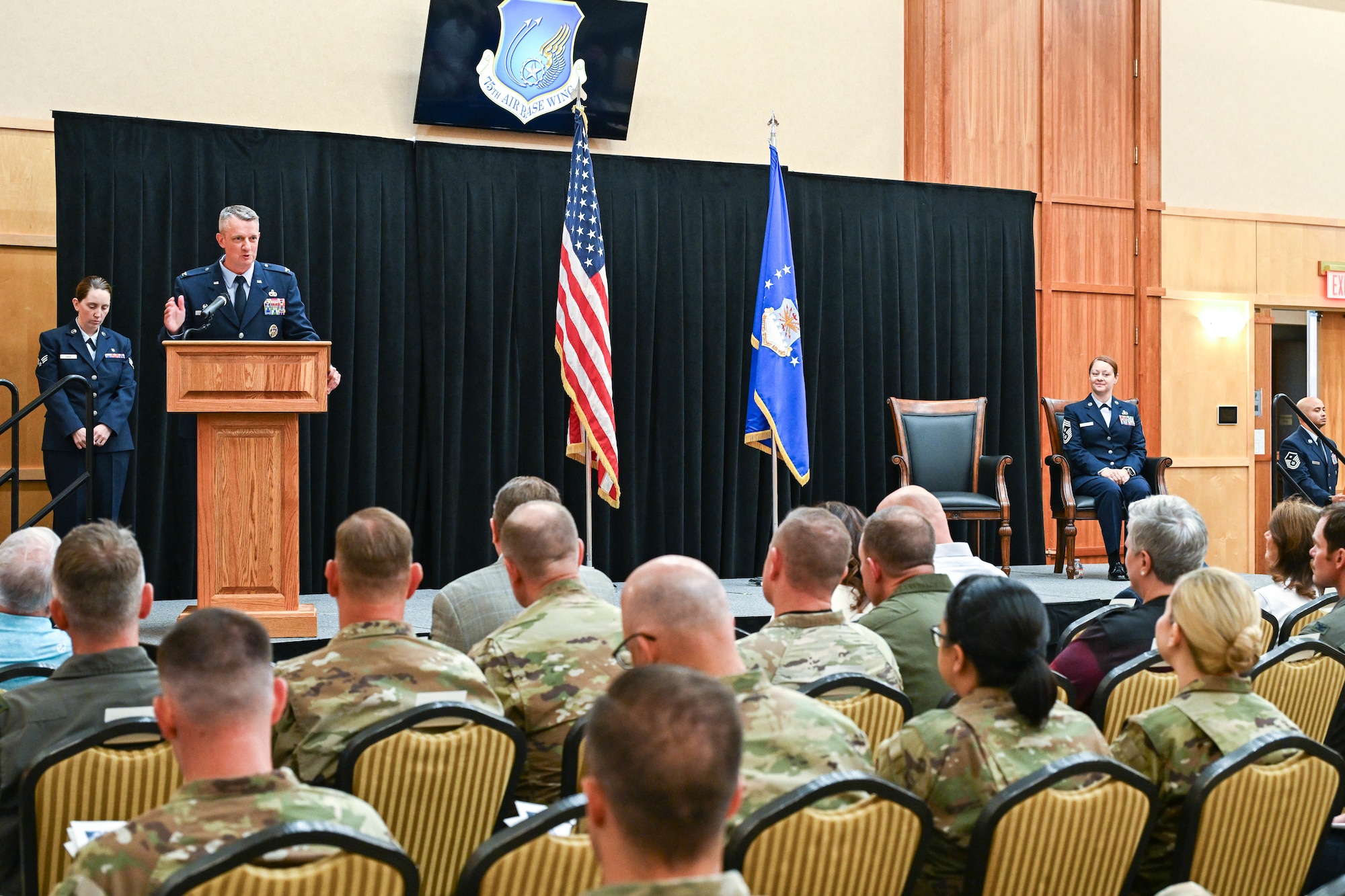 At left, Col. Cornelius delivers remarks from behind a podium in front of guests while Chief Master Sgt. Gilson sits on stage at right.