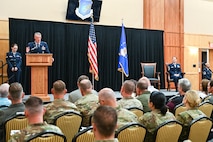 At left, Col. Cornelius delivers remarks from behind a podium in front of guests while Chief Master Sgt. Gilson sits on stage at right.