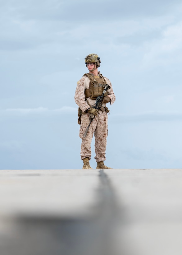 A Marine in combat gear stands on an airplane runway with a cloudy sky in the background.