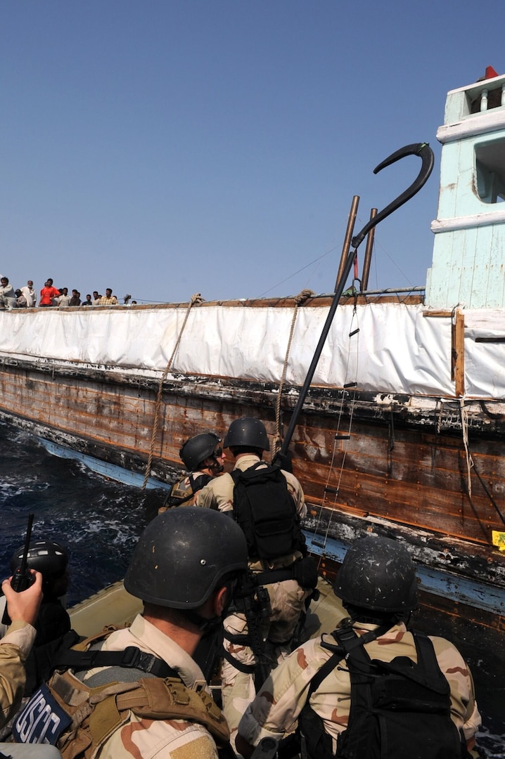 Sailors in a small boat wearing camouflage uniforms and helmets prepare to board a ship with people standing on deck.