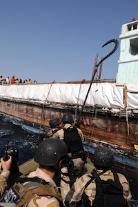 Sailors in a small boat wearing camouflage uniforms and helmets prepare to board a ship with people standing on deck.