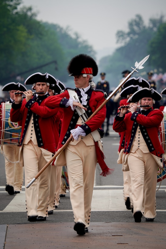 Army soldiers dressed in red Revolutionary War-era uniforms with dark tri-cornered hats are marching in formation while performing with musical instruments. The leader in front is carrying a long pike and is wearing a hat with fur down the middle.