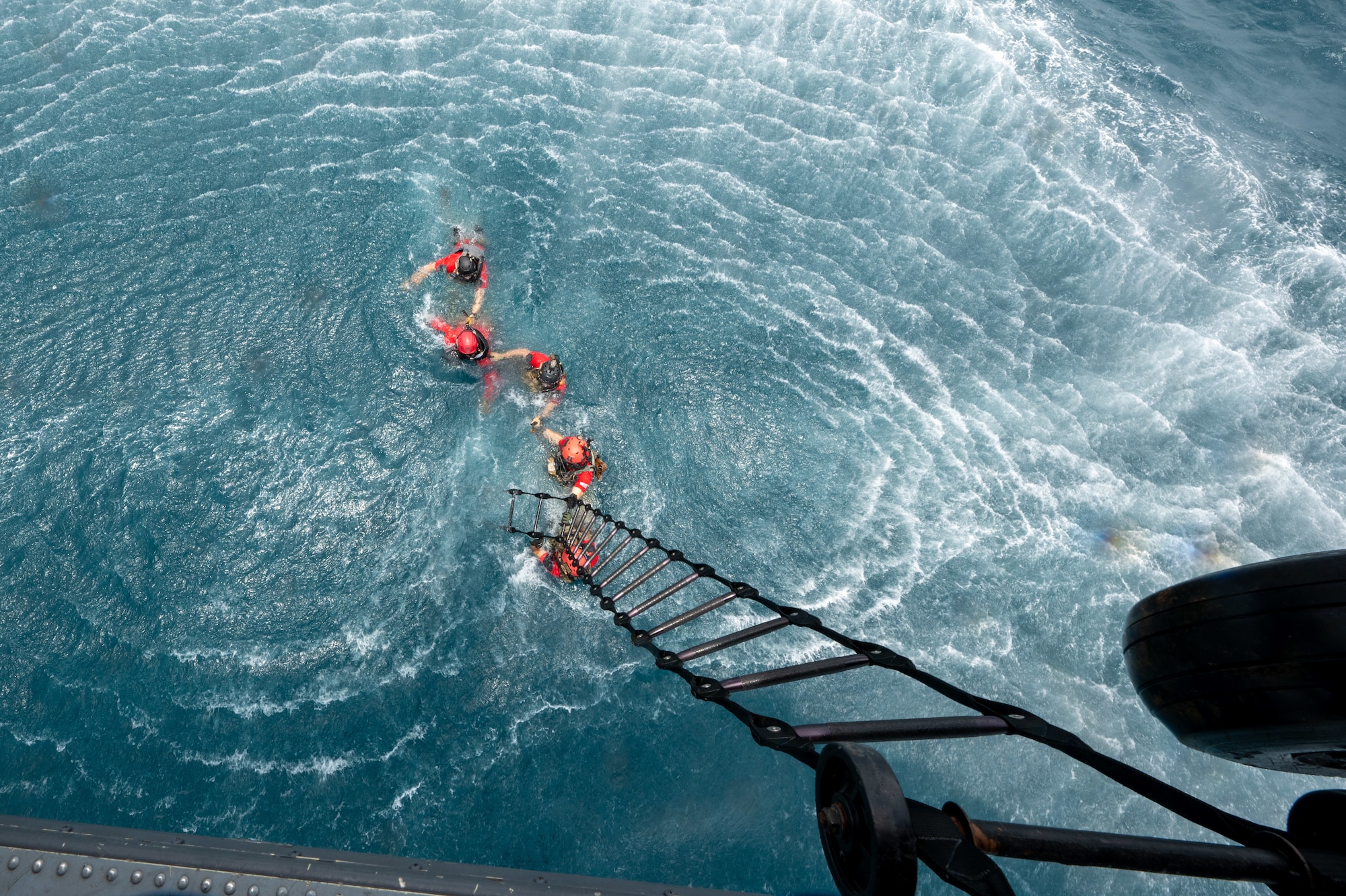 military members swim in the ocean with linked hands while reaching for a rope from a helicopter