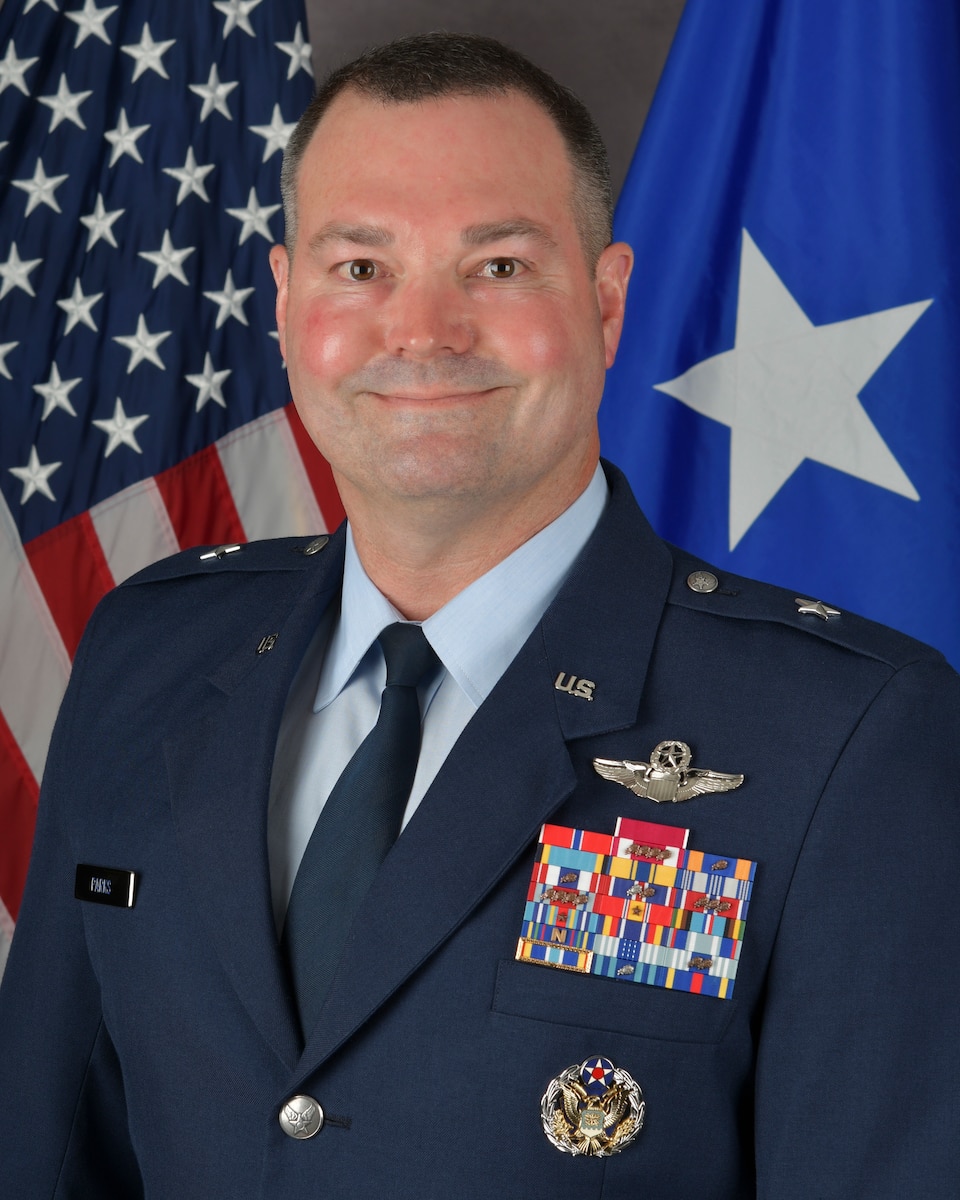 a man wearing a USAF uniform poses for a photograph in front of a US flag and one-star general officer flag