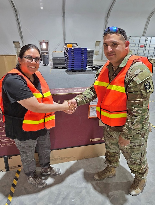 A soldier and a lady shake hands in a warehouse.