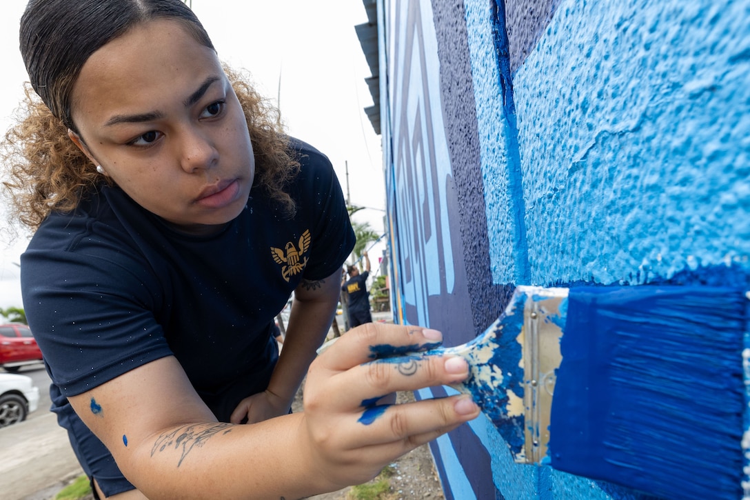 A sailor paints a blue wall outside, as seen from close-up.