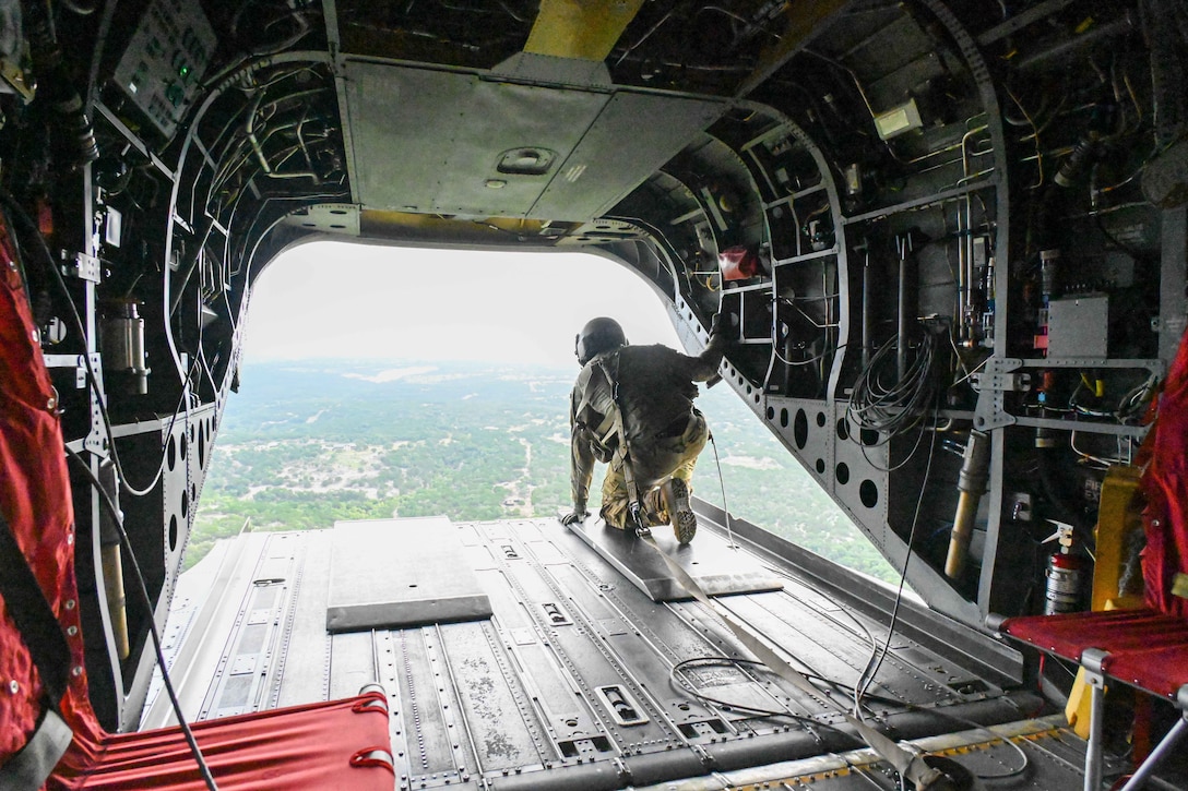 A National Guard soldier crouches on an open aircraft ramp and looks down at a forest.