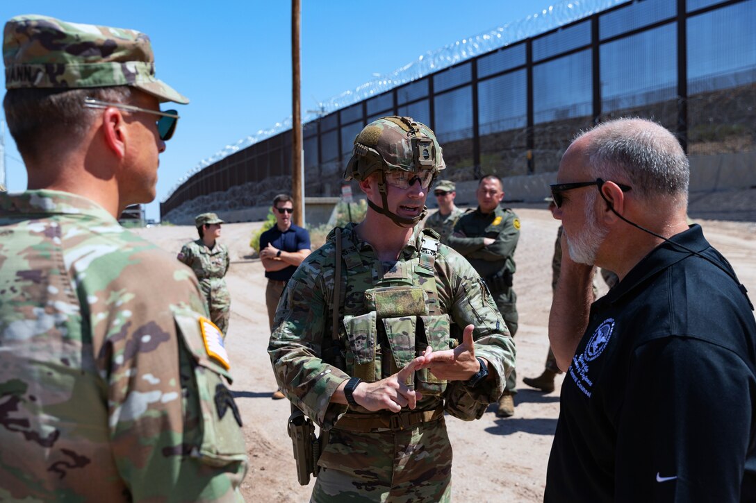 Two soldiers wearing camouflage uniforms talk to a person wearing a black, collared shirt as others stand in the background near a tall security barrier on a sunny day.