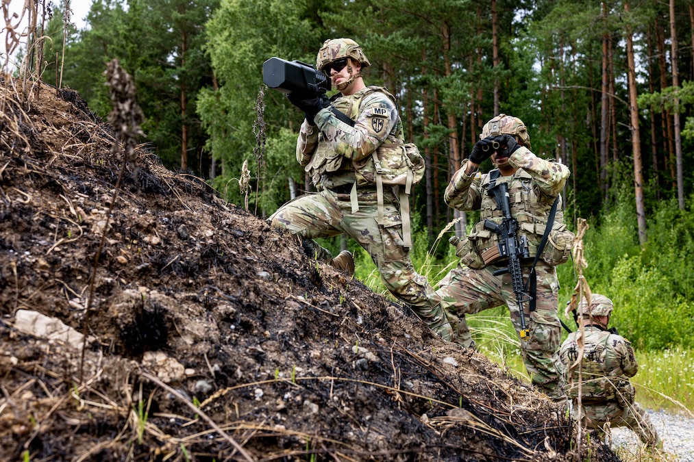 A soldier holds up military equipment on a small hill while another looks through binoculars in front of a forest.