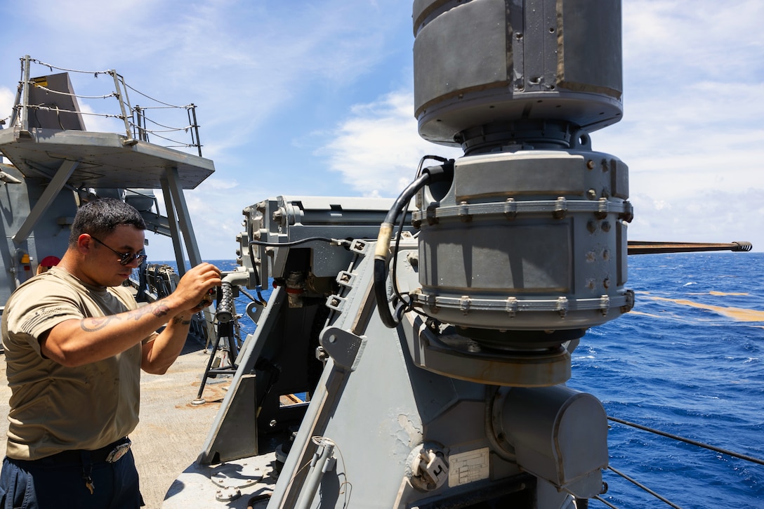 A sailor wearing a T-shirt and sunglasses works on a large gun mounted to the deck of a ship with a blue sea in the background.