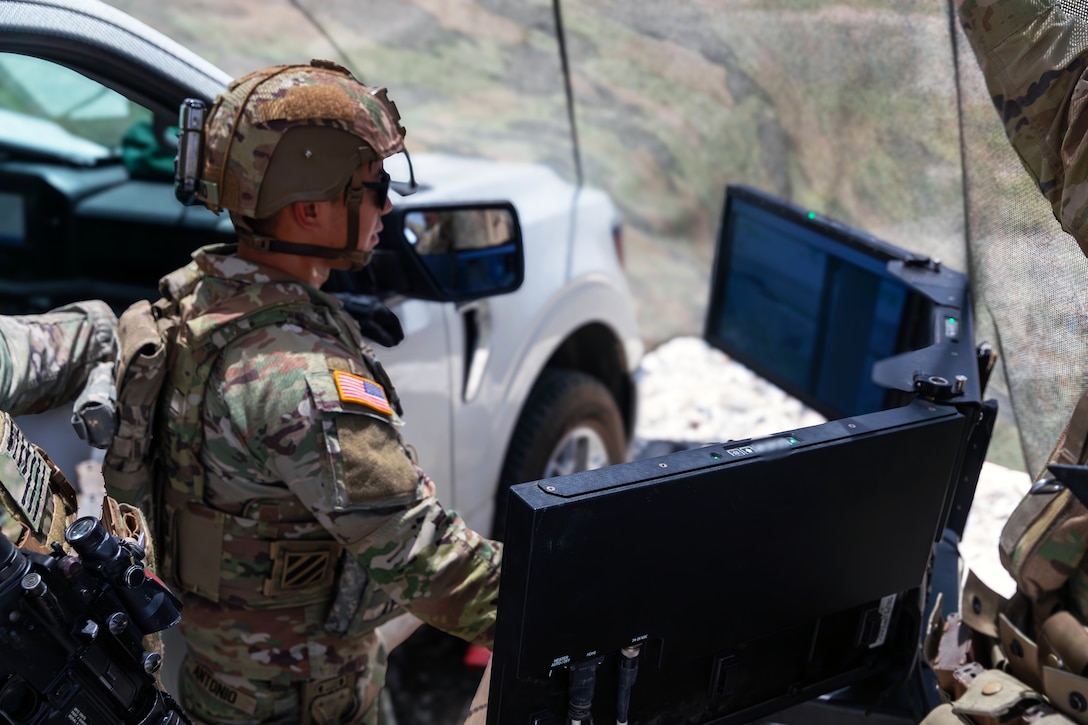 A soldier in combat gear and standing next to a white vehicle looks at electronic equipment that includes a three-screen display.