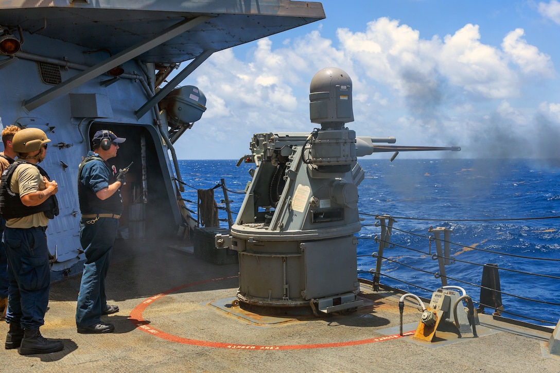 Three sailors stand behind a large gun as it is fired remotely from the deck of a ship toward a blue sea, with a cloudy, blue sky in the background.