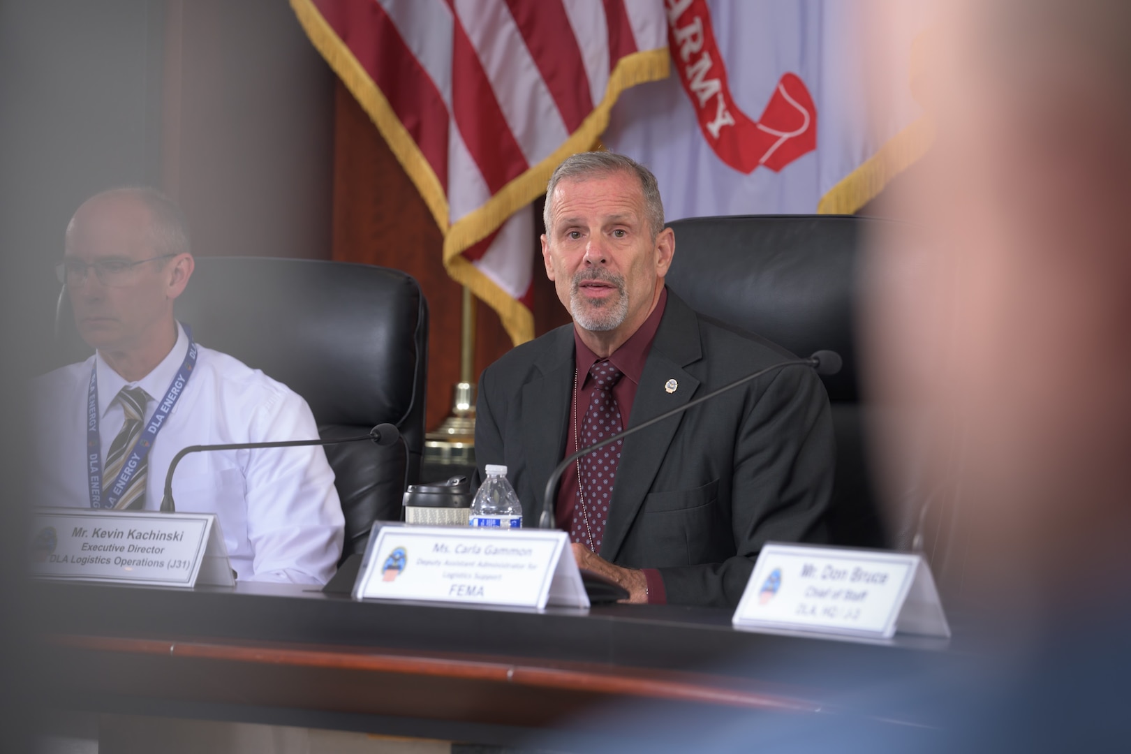 A man is seated at a conference table with flags behind him, framed in the foreground between two other participants