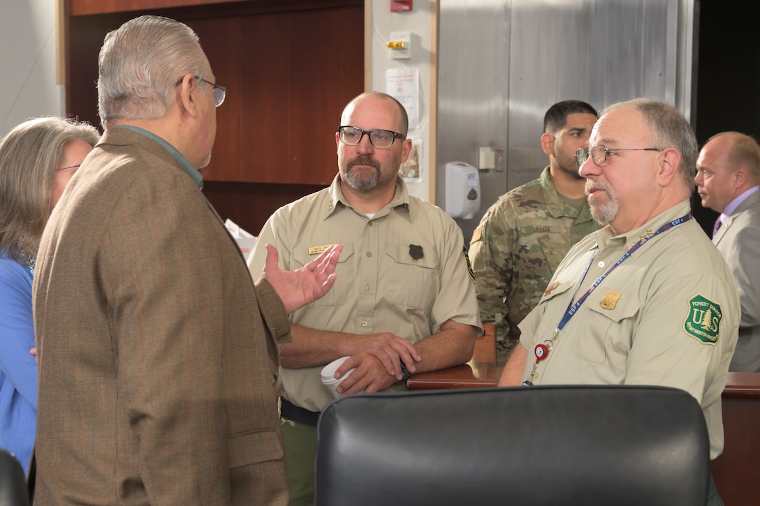 Four people are in a discussion in a conference room, with two of the people facing the camera wearing U.S. Forest Service uniforms
