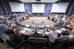 More than a dozen people sit around a C-shaped conference table facing three projector screens and a speaker behind a podium