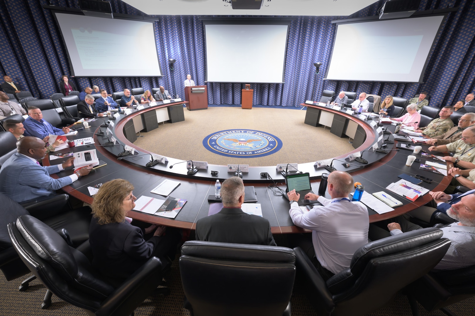 More than a dozen people sit around a C-shaped conference table facing three projector screens and a speaker behind a podium