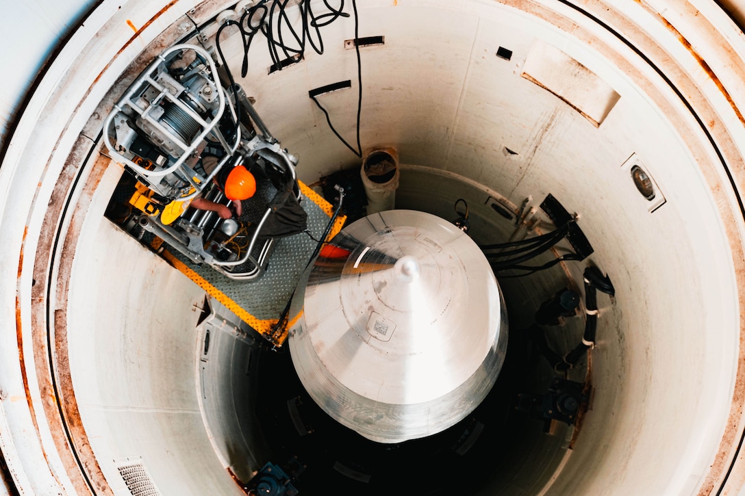 An airman in an orange construction hat stands on a platform in a launch tube, as seen from above.