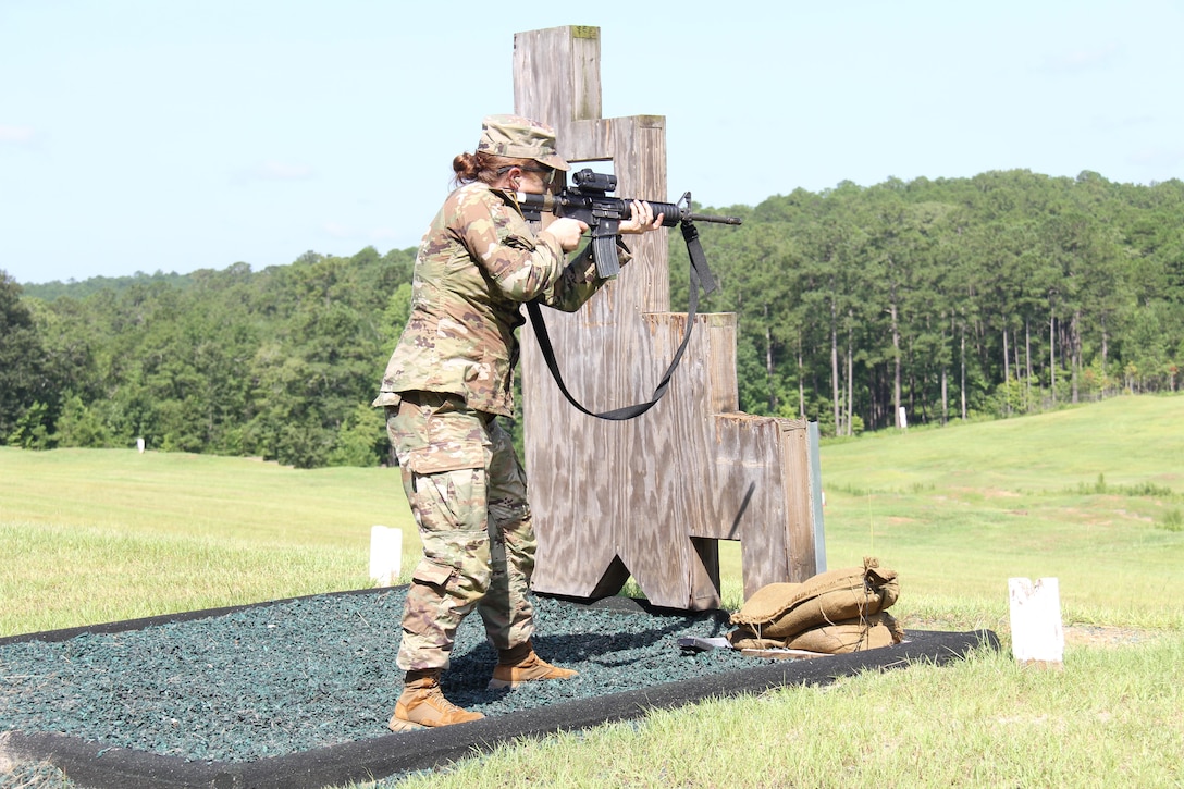 A soldier stands behind a wooden obstacle and fires a service weapon in a field.