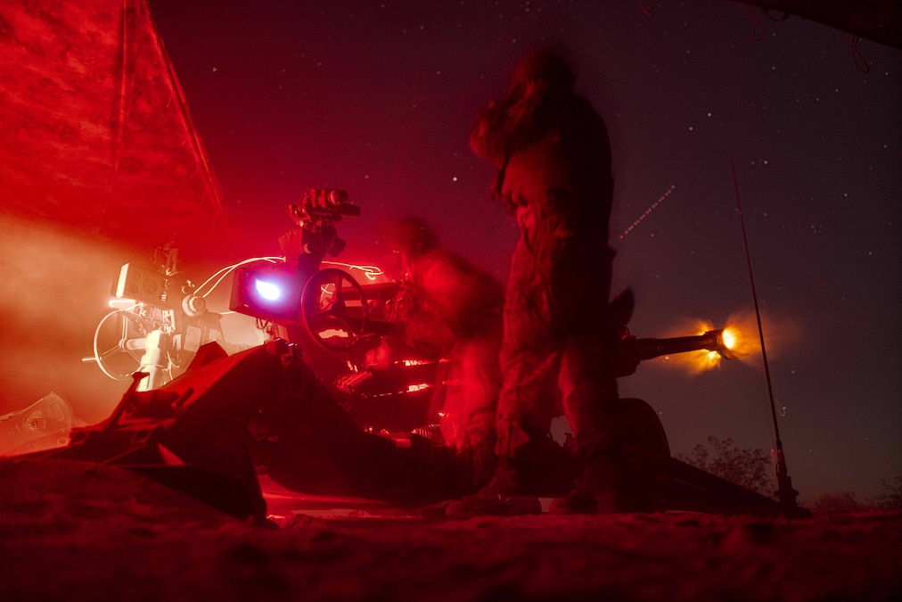 Marines fire a large weapon in the dark while surrounded by red light.