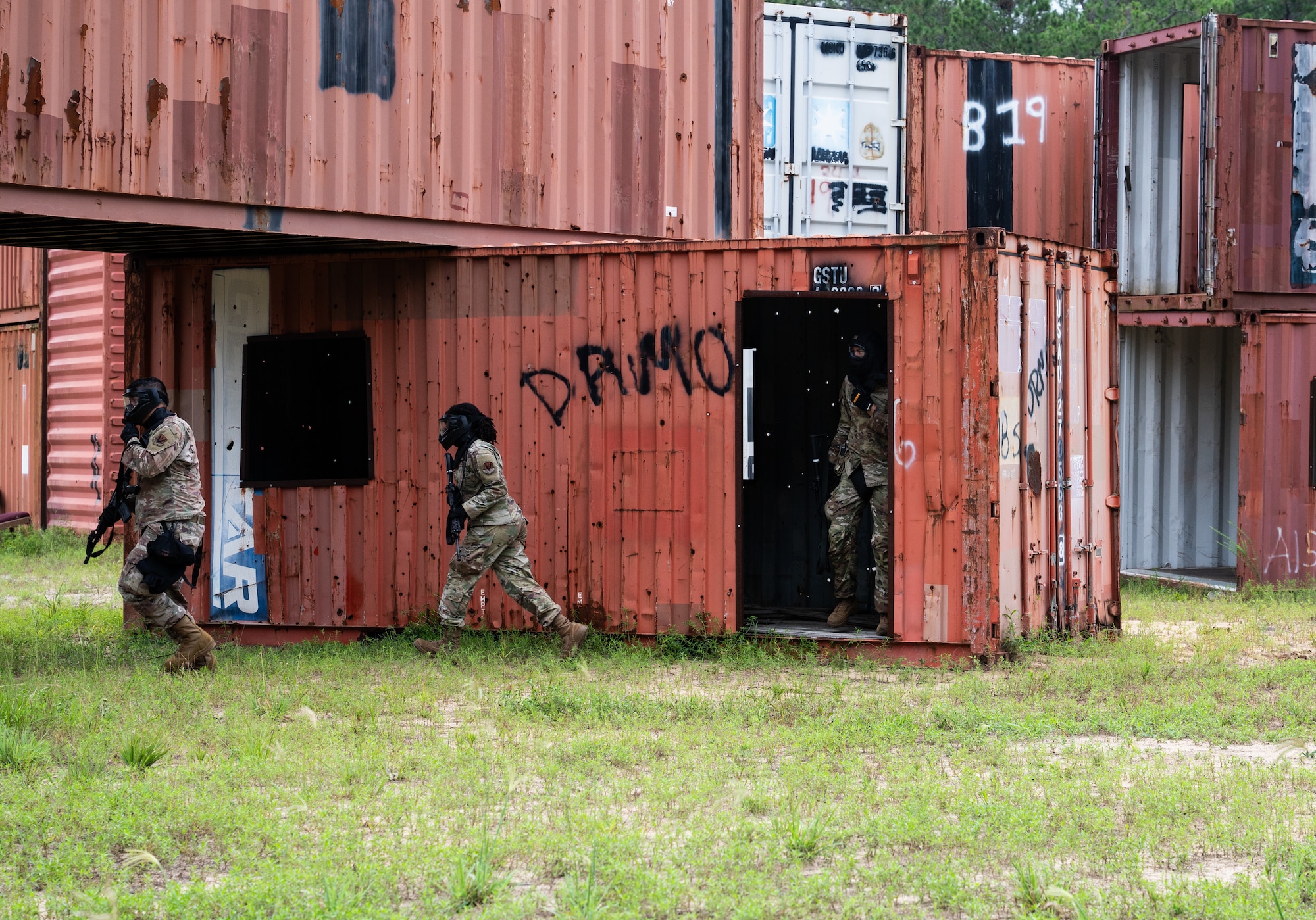 A red cargo crate is used as a defense for one uniformed member, who stands inside. Two other uniformed members run along the crate. All three members are wearing tactical masks and carrying weapons.