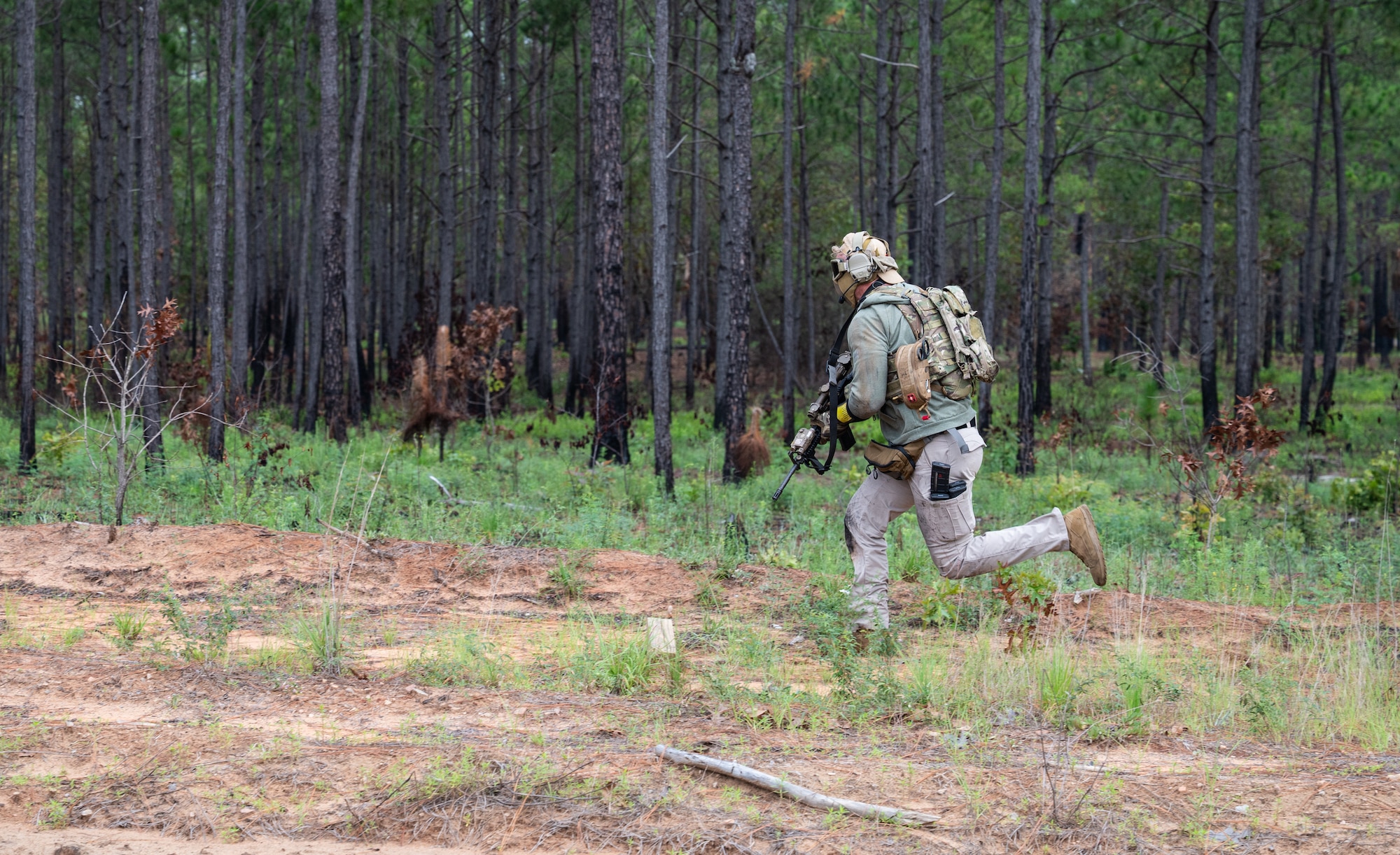 A military member in tactical gear runs along the tree line with a weapon in hand.