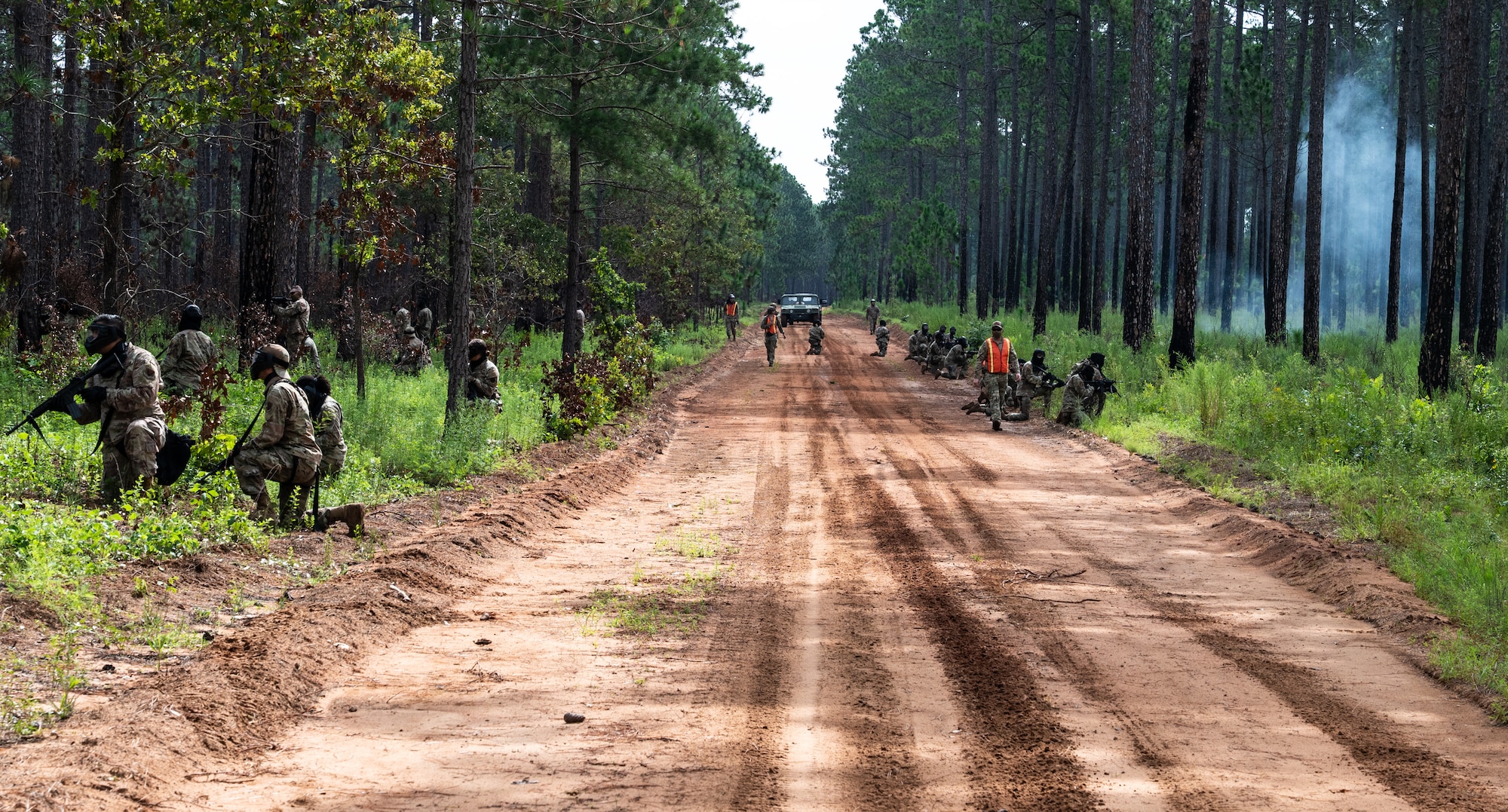 Airmen line up along the sides of a dirt road as a military vehicle comes down the middle, and a smoke bomb goes off to the right of the photo.