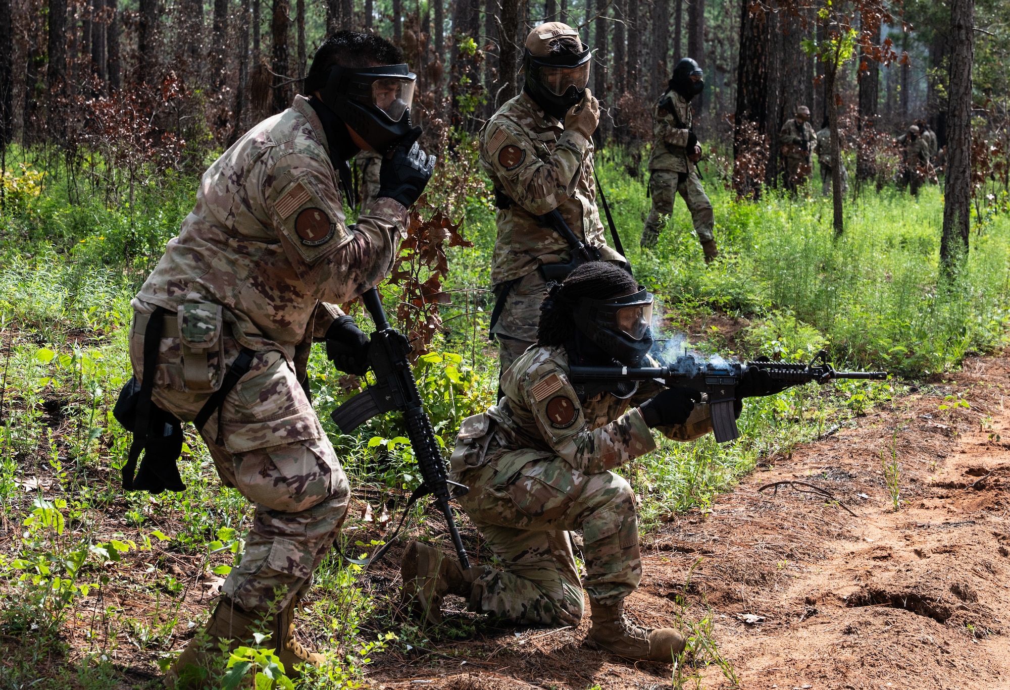 Two members in uniform watch another member aim and empty their magazine as smoke rises from the weapon.