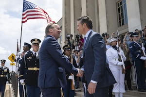 Two men in business suit shake hands in front of a building. Several men and women in military dress uniforms are standing behind them holding rifles. One service member is holding the American flag. In the background are several people holding cameras.