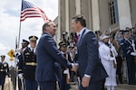 Two men in business suit shake hands in front of a building. Several men and women in military dress uniforms are standing behind them holding rifles. One service member is holding the American flag. In the background are several people holding cameras.