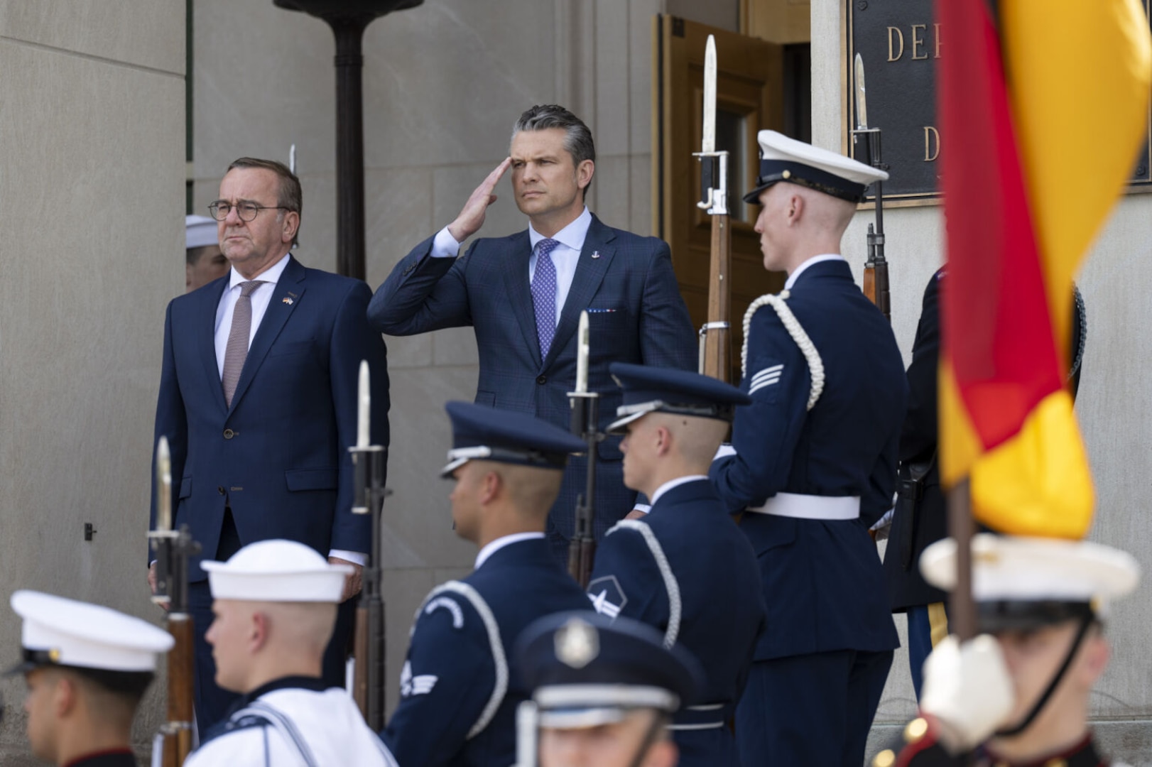 A man wearing a business suit salutes while standing next to another man in a business suite. Several military members in service dress uniforms are standing on stairs and holding rifles in front of them. One service member is holding up the German flag.