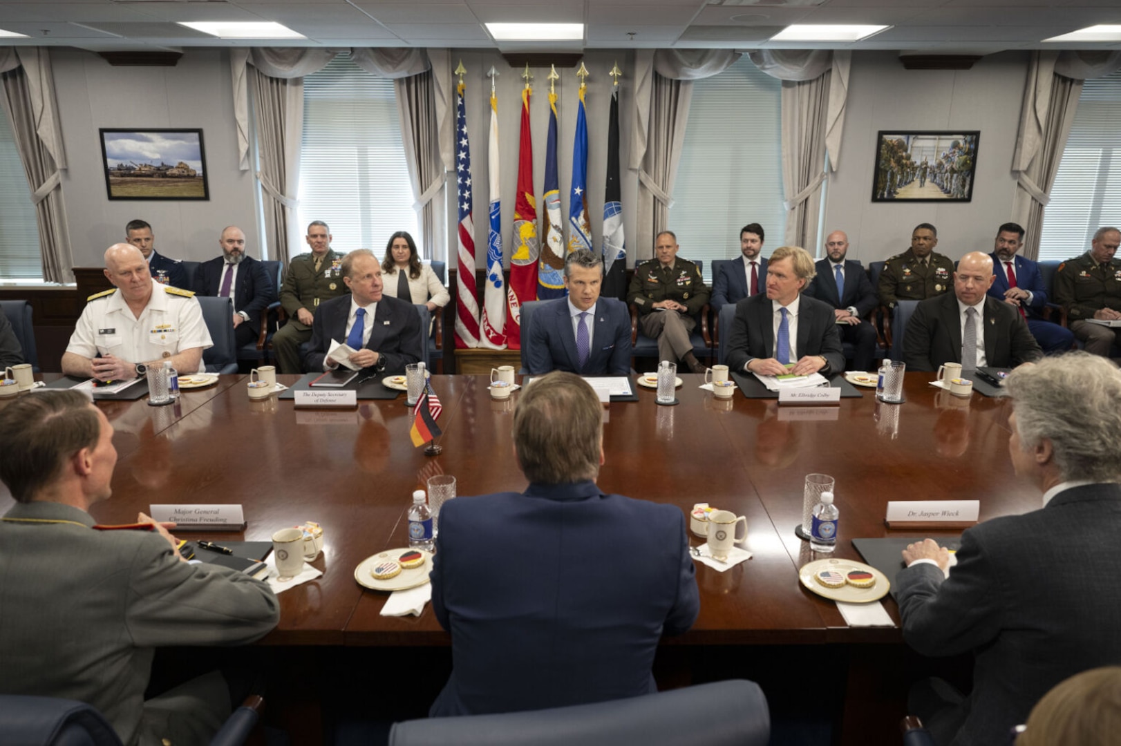 Six men in business suits and two men wearing dress military uniforms are sitting at a large wooden table; there are five men on one side and three on the opposite side. There are several men and women in business attire and military dress uniforms in the background.