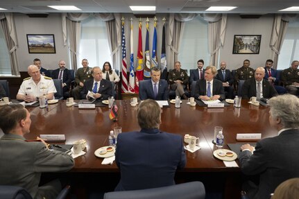 Six men in business suits and two men wearing dress military uniforms are sitting at a large wooden table; there are five men on one side and three on the opposite side. There are several men and women in business attire and military dress uniforms in the background.