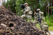 A soldier holds up military equipment on a small hill while another looks through binoculars in front of a forest.