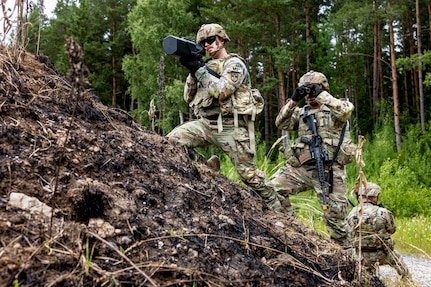 A soldier holds up military equipment on a small hill while another looks through binoculars in front of a forest.