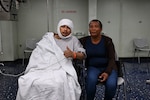 On the left a patient in a hospital gown covered in a blanket has a wrap on his head and a blood pressure machine attached to his left arm which is giving a thumbs up. On the right a woman in a blue shirt, black jacket, and jeans sits next to the patient