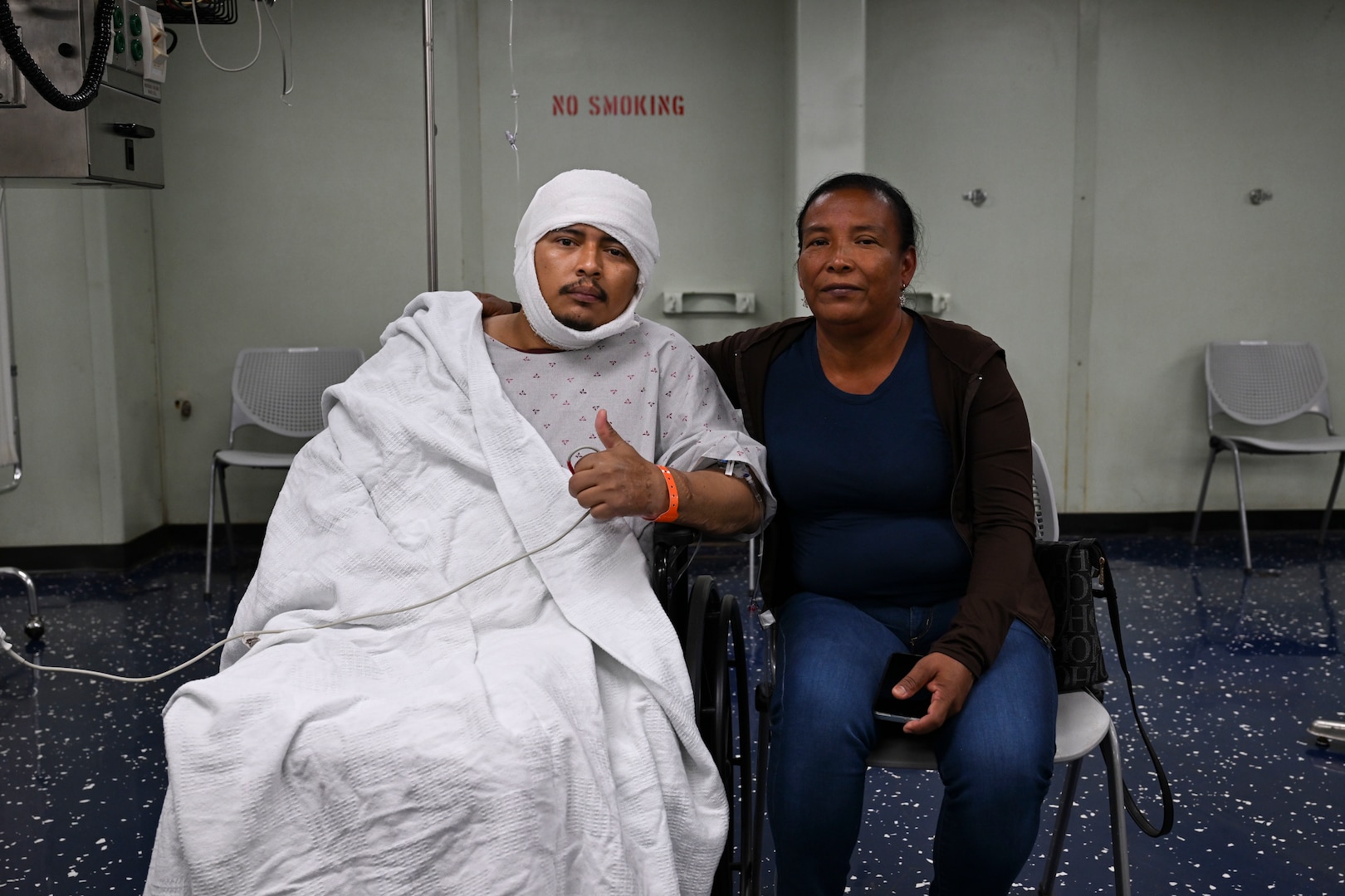 On the left a patient in a hospital gown covered in a blanket has a wrap on his head and a blood pressure machine attached to his left arm which is giving a thumbs up. On the right a woman in a blue shirt, black jacket, and jeans sits next to the patient