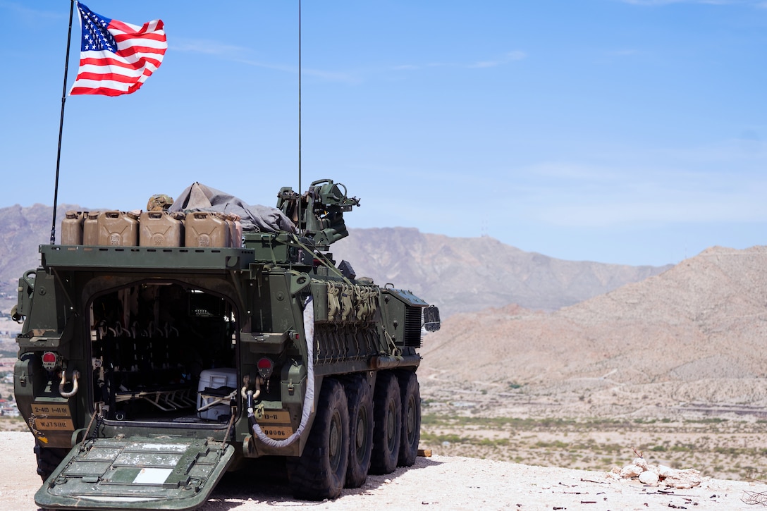 An armored vehicle flying an American flag attached to a pole is seen parked at an elevated area overlooking sparsely vegetated land with mountains in the distance.